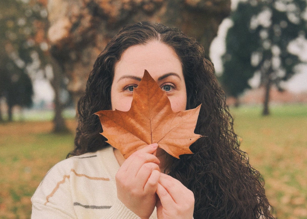 Person holding a brown leaf over their face in an outdoor setting with trees and grass.